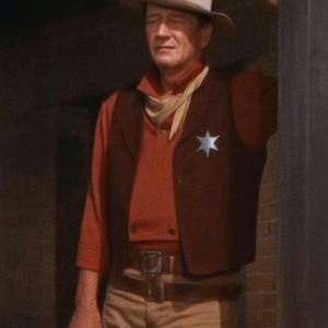 May include: A man in a cowboy outfit, wearing a light brown hat, red shirt, brown waistcoat with a star badge, and tan trousers. He is leaning against a dark doorway, holding a metal object.