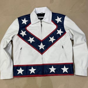 May include: White leather jacket with a star-spangled design. The jacket features a white body with navy blue and red accents, and white stars. It has a zipper closure and two front pockets. The collar is white.