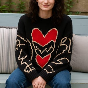 Puede incluir: Suéter negro con un gran diseño de corazón rojo con contorno blanco. El suéter tiene texto blanco en las mangas. La persona lleva vaqueros azules y está sentada en un banco azul con cojines a rayas.