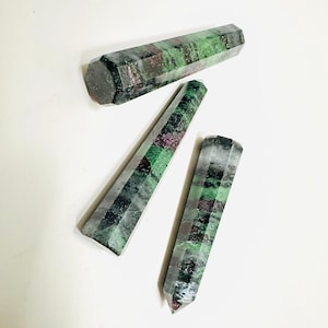 May include: Three polished gemstone wands. The wands are a mix of green, red, and grey colours. The wands have a hexagonal shape and are tapered at one end. The wands are arranged on a white surface.