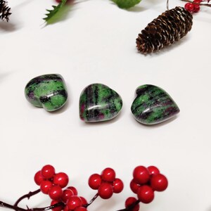 Op de afbeelding: Drie groene en roze hartvormige stenen op een witte achtergrond. De stenen worden omringd door rode bessen en een dennenappel.