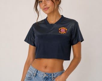 May include: A navy blue short-sleeved crop top with a V-neckline. The shirt features a graphic on the upper left chest consisting of a pink pair of lips surrounded by the orange text "POWER LOVE" and small orange hearts.