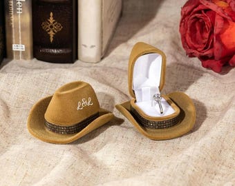 May include: Two brown velvet cowboy hats, one with a ring box and the other with the initials "L&L" embroidered on the band. The ring box is open and a diamond ring is inside.