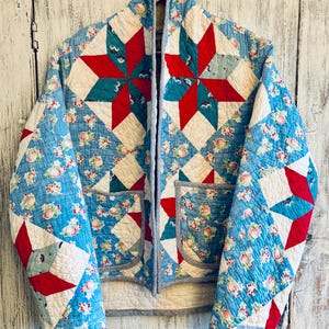 May include: A quilted jacket featuring a star pattern in red, white, and teal. The fabric has a light blue background with a floral print of small pink and green flowers. The jacket includes two front pockets and a grey trim along the edges.