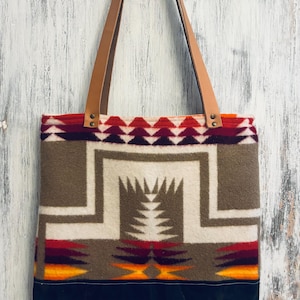 May include: A tote bag featuring a geometric pattern in shades of brown, cream, red, orange, and purple. The upper section has a patterned wool-like texture, while the base is solid black. It includes two light brown leather shoulder straps.