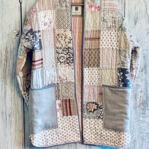 May include: A long-sleeved quilted jacket featuring a patchwork design of various floral, striped, and geometric patterns in muted tones of pink, beige, grey, and white. The jacket includes two large solid grey patch pockets and a label at the neck.