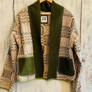 May include: A patterned jacket with olive green details hangs on a wooden hanger. The jacket has a mix of beige, brown, and pink woven fabric with a geometric design. The collar and pockets are a solid olive green.