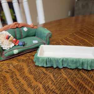 K&ouml;nnte beinhalten: Ein gr&uuml;nes Miniatursofa mit einem dekorativen Kissen und einer kleinen Figur einer Person in roter und blauer Kleidung. Daneben befindet sich eine wei&szlig;e rechteckige Schale mit gr&uuml;nem R&uuml;schenbesatz. Die Gegenst&auml;nde befinden sich auf einer Holzoberfl&auml;che.