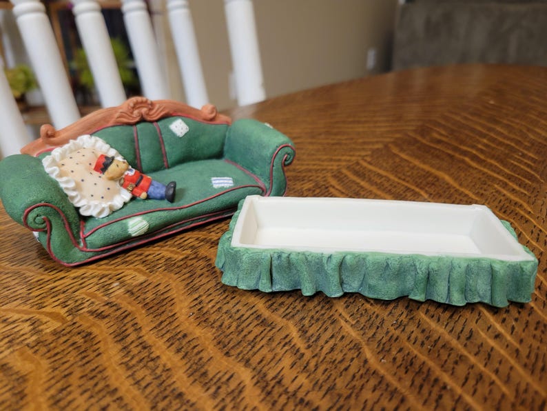 K&ouml;nnte beinhalten: Ein gr&uuml;nes Miniatursofa mit einem dekorativen Kissen und einer kleinen Figur einer Person in roter und blauer Kleidung. Daneben befindet sich eine wei&szlig;e rechteckige Schale mit gr&uuml;nem R&uuml;schenbesatz. Die Gegenst&auml;nde befinden sich auf einer Holzoberfl&auml;che.