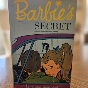 May include: Vintage hardcover book titled "Barbie's Secret" with a light blue cover and yellow lettering. The cover illustration depicts Barbie and a boy in a car, with a house in the background. A Random House Book.