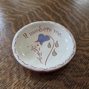 Puede incluir: Pequeño plato de cerámica blanquecina con un diseño floral y el texto "It wonders me". El plato presenta una flor azul, tallos marrones y formas de lágrima verdes. El borde está delineado en color marrón rojizo.