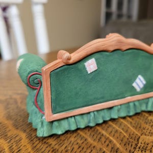 K&ouml;nnte beinhalten: Ein gr&uuml;nes Miniatursofa mit Holzrahmen und gr&uuml;nem Kissen. Das Sofa hat einen ger&uuml;schten Rock und dekorative Aufn&auml;her auf der R&uuml;ckseite. Das Sofa ist ein dekorativer Gegenstand.