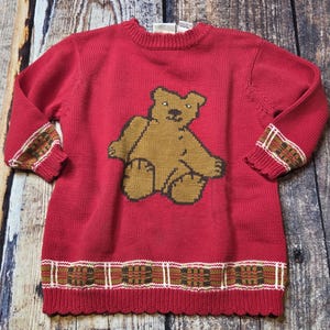 May include: A red knit sweater featuring a brown bear design on the front. The sweater has decorative trim on the cuffs and hemline with a repeating pattern of brown, white, and green. The sweater is made of a soft, comfortable material.