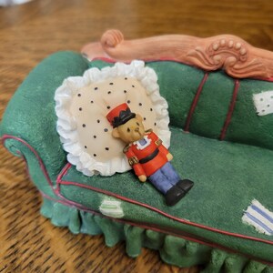 K&ouml;nnte beinhalten: Ein dekoratives Miniatursofa in Gr&uuml;n mit roten Zierleisten, einem wei&szlig;en Kissen mit schwarzen Punkten und einer kleinen Spielzeugsoldatenfigur. Das Sofa hat einen ger&uuml;schten Rock und eine detaillierte Holzlehne.