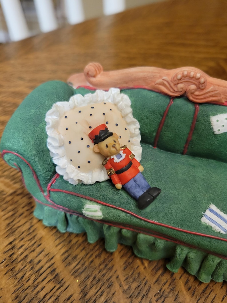 K&ouml;nnte beinhalten: Ein dekoratives Miniatursofa in Gr&uuml;n mit roten Zierleisten, einem wei&szlig;en Kissen mit schwarzen Punkten und einer kleinen Spielzeugsoldatenfigur. Das Sofa hat einen ger&uuml;schten Rock und eine detaillierte Holzlehne.