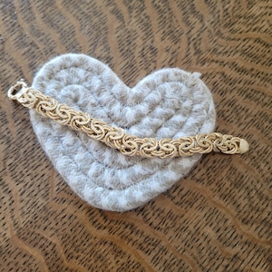 May include: A gold chain bracelet with a woven design. The bracelet is on a white heart-shaped surface.