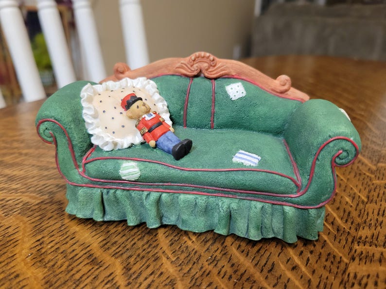 K&ouml;nnte beinhalten: Ein gr&uuml;nes Miniatursofa mit dekorativem Holzrahmen und roter Zierleiste. Eine kleine Spielzeugsoldatenfigur liegt auf einem gepunkteten Kissen. Das Sofa hat einen ger&uuml;schten Rock und dekorative Patches.