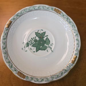 May include: White ceramic saucer with a scalloped edge, featuring a green floral design in the center and a green and gold border. The saucer has a delicate, ornate pattern and is set against a wooden surface.