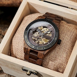 Może przedstawiać: Drewniany zegarek na rękę z brązowym paskiem i ciemną tarczą, prezentowany w drewnianym pudełku. Tarcza zegarka odsłania wewnętrzny mechanizm ze złotymi kołami zębatymi. Pudełko jest jasne z metalowym zapięciem.