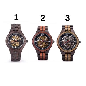 May include: Three wooden watches with visible internal mechanisms are displayed. The watches have dark brown, reddish-brown, and light brown wooden bands and cases. Each watch face features the number 12 at the top and a detailed view of the inner workings.