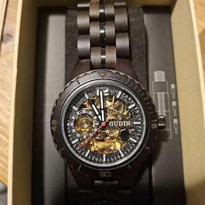 May include: A dark brown wooden watch with a black face and band, presented in a cardboard box. The watch face features gold-colored gears and the brand name "GUDIN". The watch band is made of interlocking wooden links.
