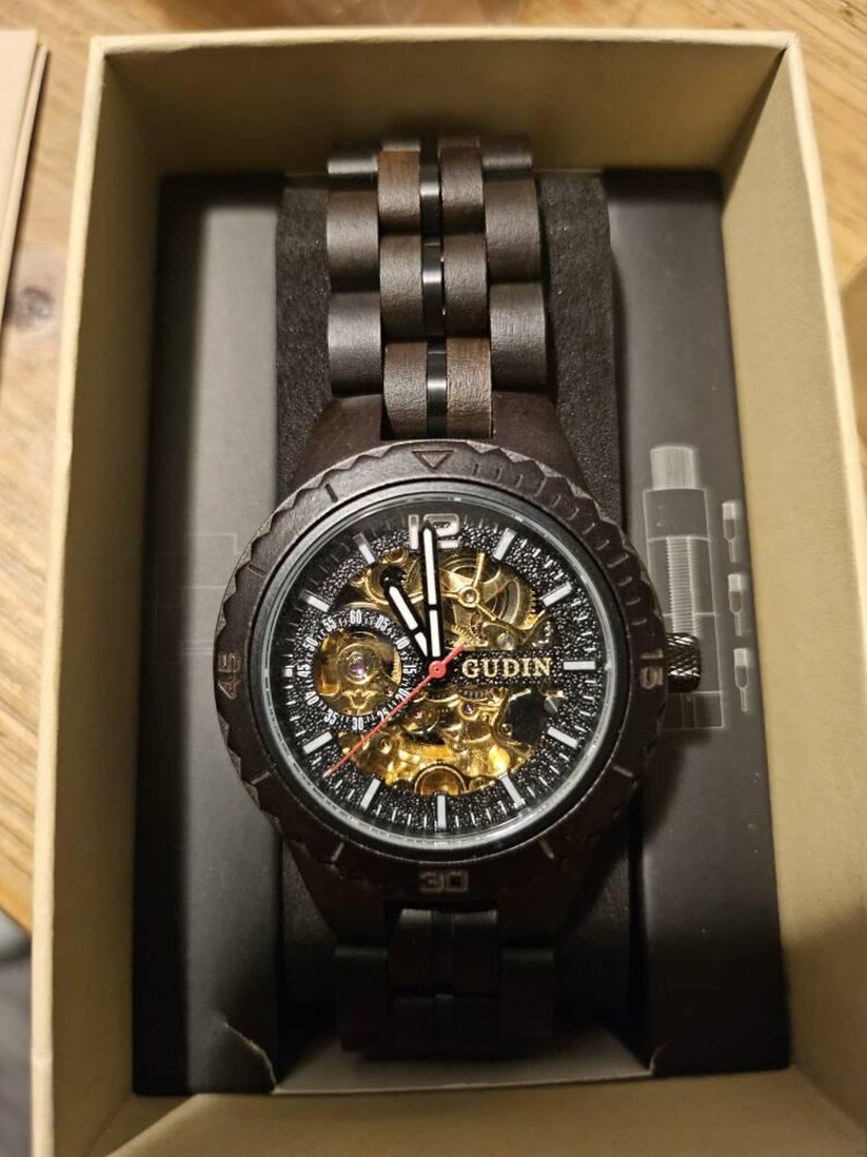 May include: A dark brown wooden watch with a black face and band, presented in a cardboard box. The watch face features gold-colored gears and the brand name "GUDIN". The watch band is made of interlocking wooden links.