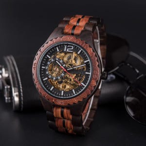 Puede incluir: Un reloj de pulsera de madera con esfera oscura y engranajes visibles. El reloj presenta un diseño de veta de madera marrón y negra en la correa y el bisel. La esfera del reloj muestra el número 12 de forma destacada.