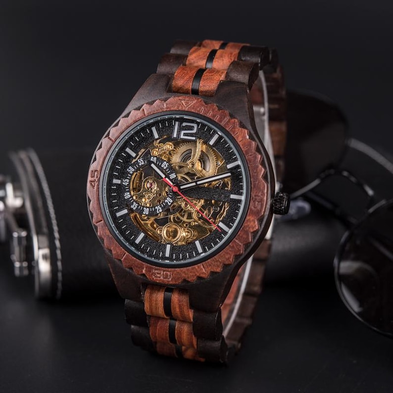 May include: A wooden wristwatch with a dark face and visible gears. The watch features a brown and black wood grain design on the band and bezel. The watch face displays the number 12 prominently.