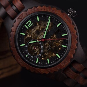 May include: A wooden wristwatch with a black face and a visible mechanical movement. The watch has a dark brown wooden bezel and band. The watch hands and hour markers glow green. The watch face displays the numbers 12, 15, 20, 25, 30, 35, 40, 45, 50, and 55.