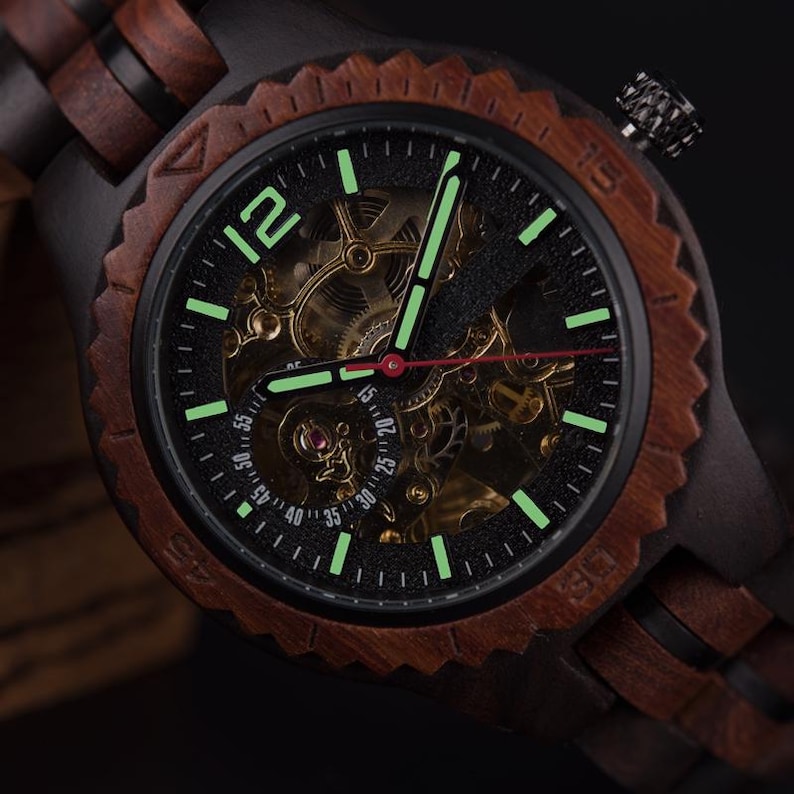 May include: A wooden wristwatch with a black face and a visible mechanical movement. The watch has a dark brown wooden bezel and band. The watch hands and hour markers glow green. The watch face displays the numbers 12, 15, 20, 25, 30, 35, 40, 45, 50, and 55.
