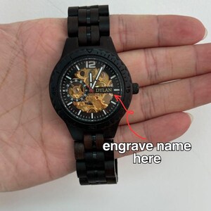 May include: Black wooden wristwatch with a visible gold-toned mechanical movement. The watch face displays the name "DYLAN" and the number "12". The watch has a black wooden band and a black bezel. The text "engrave name here" is also visible.