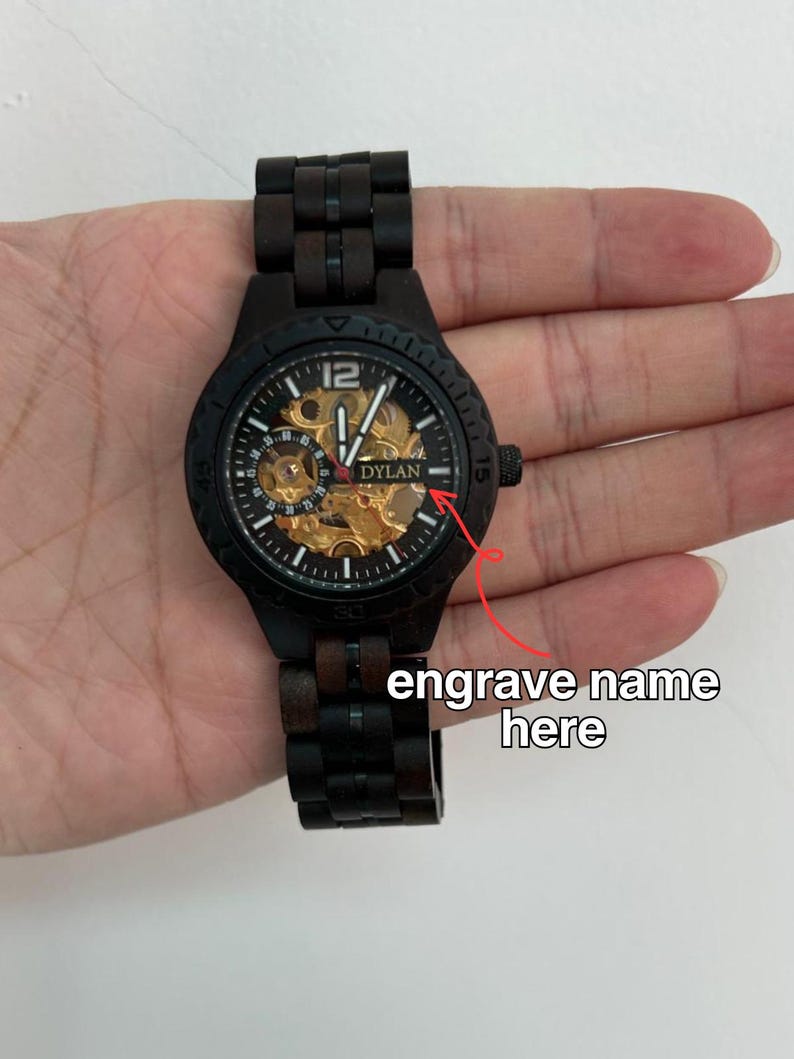 May include: Black wooden wristwatch with a visible gold-toned mechanical movement. The watch face displays the name "DYLAN" and the number "12". The watch has a black wooden band and a black bezel. The text "engrave name here" is also visible.