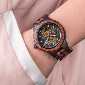 May include: A dark brown wooden watch with a black face and visible gears. The watch has a brown wooden bezel and band. The watch is on a wrist, and the background is a light pink fabric.
