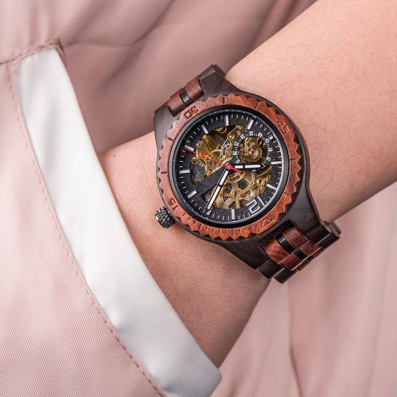 May include: A dark brown wooden watch with a black face and visible gears. The watch has a brown wooden bezel and band. The watch is on a wrist, and the background is a light pink fabric.