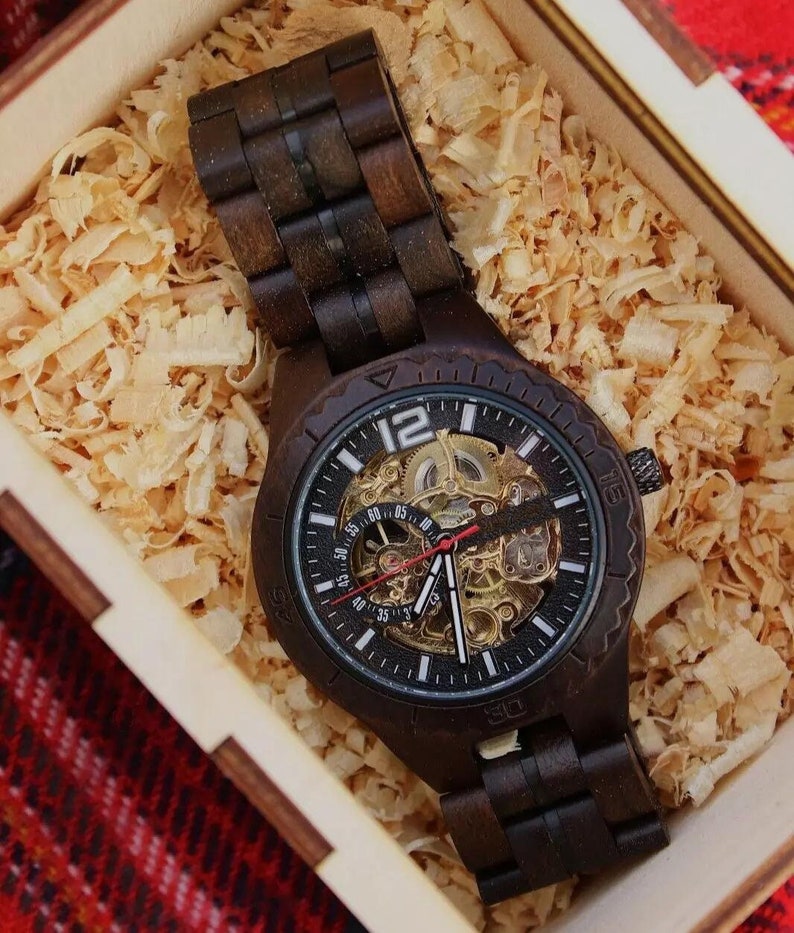 May include: A dark brown wooden watch with a black face and visible inner workings. The watch is displayed in a wooden box filled with wood shavings. The watch has a matching wooden band.