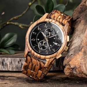 May include: A wooden wristwatch with a black face and gold-colored hands and accents. The watchband and watch case are made of wood with a striped pattern. The watch is displayed on a wooden surface.