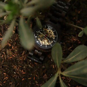 May include: A dark wooden wristwatch with a black face and gold-colored internal gears. The watch has a visible mechanical movement and a dark wooden band. The watch is surrounded by green foliage and brown earth.