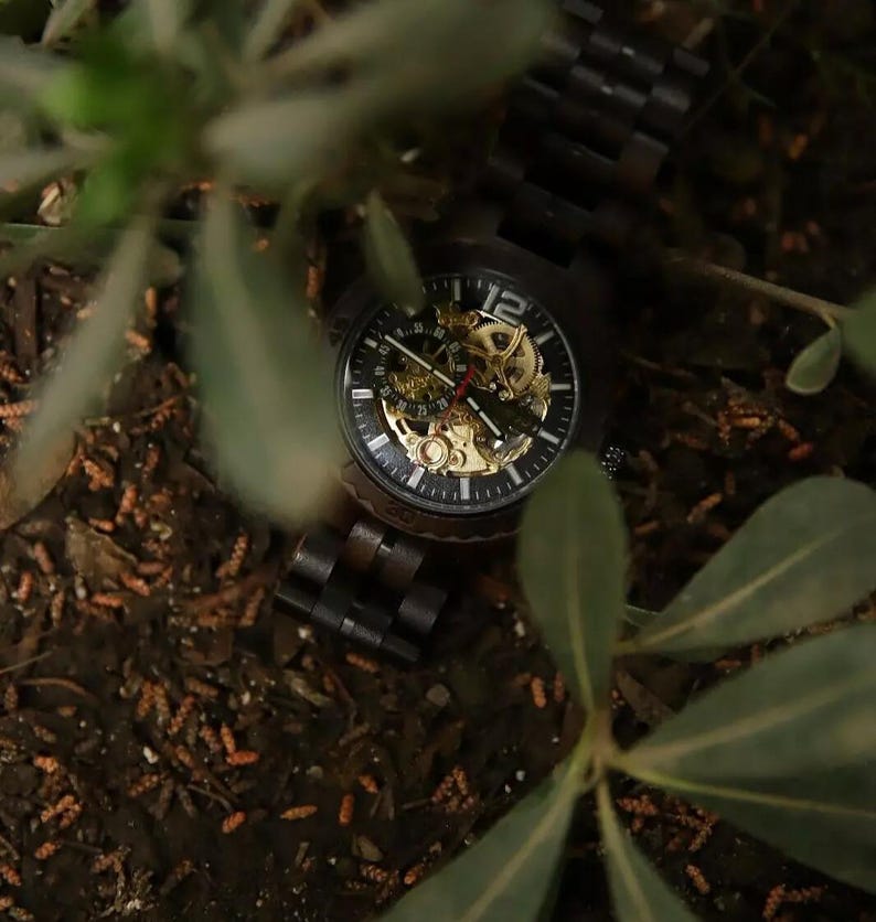 May include: A dark wooden wristwatch with a black face and gold-colored internal gears. The watch has a visible mechanical movement and a dark wooden band. The watch is surrounded by green foliage and brown earth.