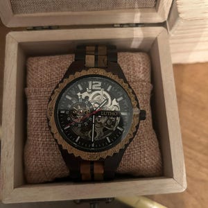 May include: A wooden wristwatch with a black face and visible gears, presented in a wooden box. The watch features a brown and black wooden bezel and band. The watch face displays the brand name "LUTHO".