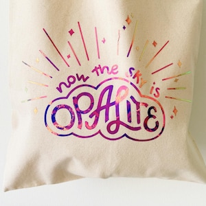 Pode incluir: Bolsa tote de lona bege com o texto "now the sky is OPALITE" em um design colorido e iridescente. O design inclui uma forma de nuvem e linhas radiantes. A bolsa tem duas alças e é adequada para transportar objetos.