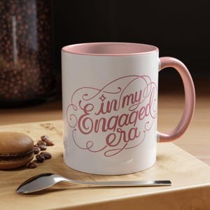 Puede incluir: Taza de cerámica blanca con interior y asa rosas. La taza presenta la frase "in my engaged era" en una escritura rosa decorativa. Una cuchara plateada y un macaron están sobre una superficie de madera.