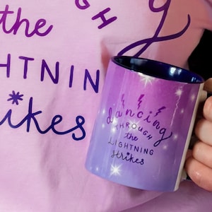 Könnte beinhalten: Eine weiße Keramiktasse mit einem Farbverlauf in Lila und Blau, auf der "dancing through the lightning strikes" steht. Die Tasse wird in einer Hand gehalten, im Hintergrund ist ein rosa Oberteil zu sehen.