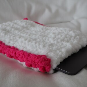 May include: White crocheted laptop sleeve with a pink crocheted border. The sleeve is lying on a white surface with a laptop inside.