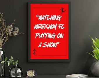 Può includere: Una stampa incorniciata rossa con testo bianco che recita "Watching Wrexham FC Putting on a Show". Il testo è in stile calligrafico. Ci sono due silhouette nere di calciatori sulla stampa.