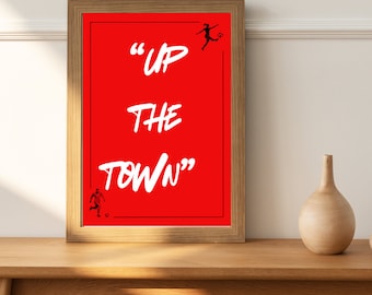 Può includere: Una stampa incorniciata con sfondo rosso e testo bianco che recita "UP THE TOWN". Ci sono due silhouette nere di persone che giocano a calcio sulla stampa.