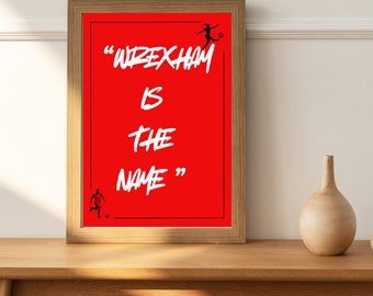 Può includere: Una stampa incorniciata con sfondo rosso e testo bianco che recita "Wrexham is the name". Ci sono due silhouette nere di calciatori sulla stampa.