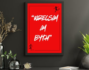 Può includere: Una stampa incorniciata con sfondo rosso e testo bianco che recita "Wrexham am Byth". Ci sono due silhouette nere di persone che giocano a calcio sulla stampa.
