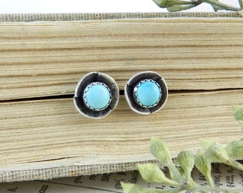 May include: A pair of silver stud earrings with turquoise stones set in a circular bezel. The earrings are on a book with a light brown cover.