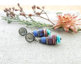 May include: A pair of handcrafted earrings with silver-tone studs and dangling beaded accents. The earrings feature blue, turquoise, and mauve beads, with a mix of textures and patterns. The earrings are displayed on a textured, neutral-colored surface.
