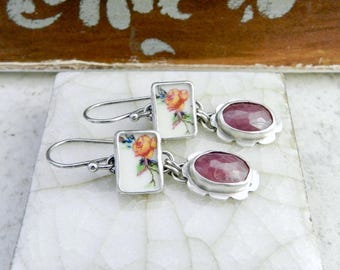 May include: Silver earrings with a rectangular floral design and oval, pink gemstones. The rectangular portion features a rose illustration in shades of orange, yellow, and green. The earrings are displayed on a white, marble-like surface.
